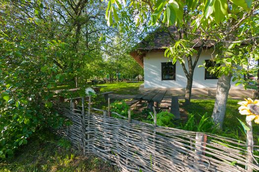 A house with white, whitewashed walls thatched roof and neat windows standing in a courtyard on a green grass fenced with a wooden fence.