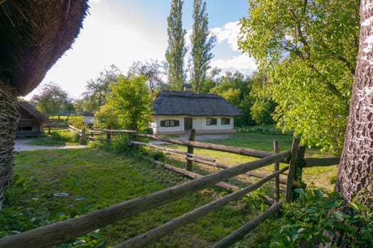 An old rural house with thatched roof standing in a courtyard fenced with wooden long poles.