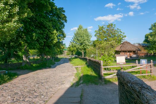 An ancient rural street with a road paved with cobblestones with wooden houses standing in courtyards fenced with a wattle fence with tall trees growing near them.