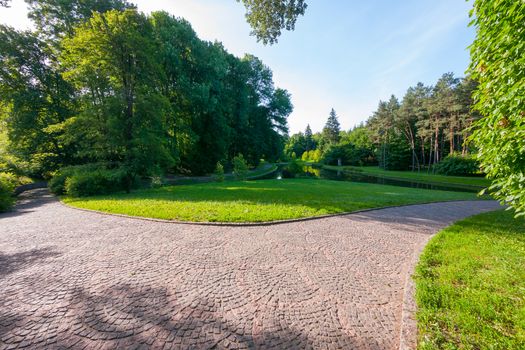 The path is laid out with a stone pavement in a beautiful green park on a summer cloudy day. A good place to relax by the lake.