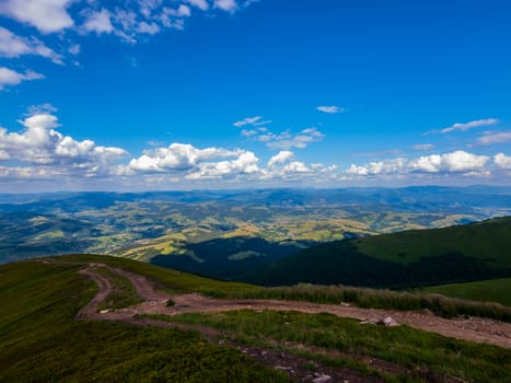 A magnificent landscape of mountains and valleys with a dirt road going down from a green slope and low floating clouds over all this beauty.