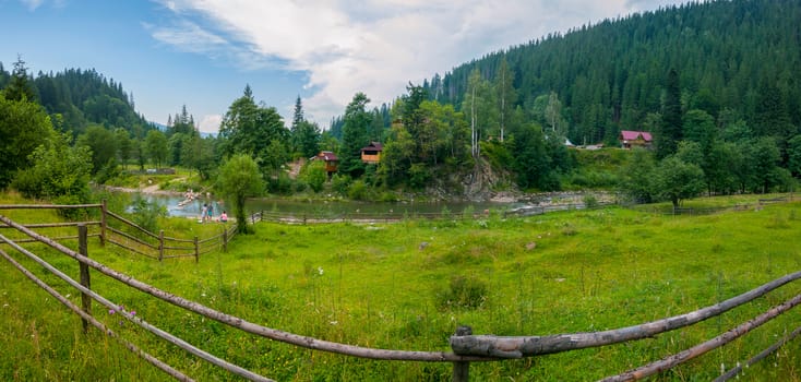 Beautiful views of the mountainous terrain with the current river between the low banks with green grass on one side and trees and houses on the other.