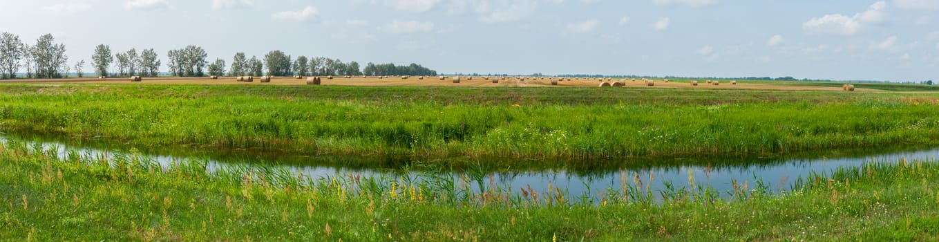A small river channel against a background of green grass and a golden field with haystacks in the distance