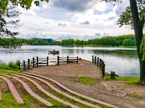 a path to the pier on the lake with a boat, a cloudy sky and green trees around. resting place, fishing