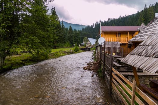 A stormy river washes the banks with a wooden house against the background of the Carpathian mountains under the cloudy sky