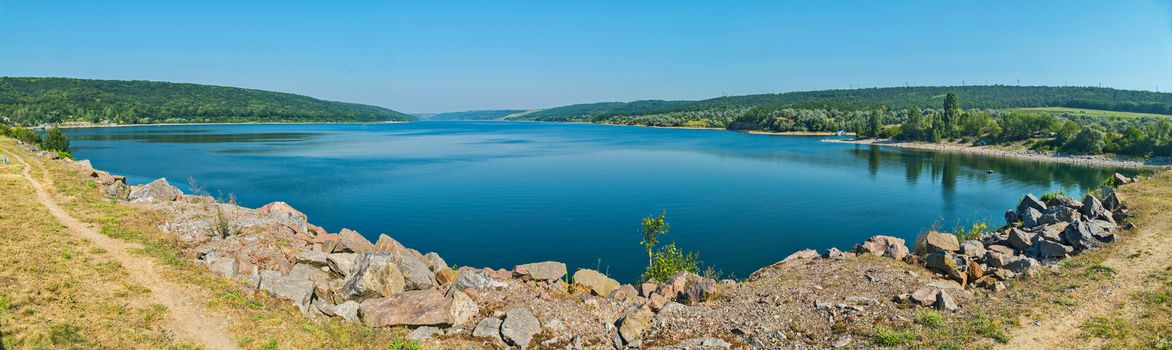 Stone shore at the blue clear lake against the background of green mountains and sky