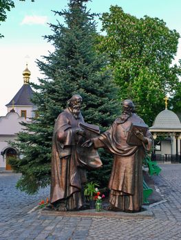 Sculpture of two saints standing near the elegant green fir tree on the background of a small chapel and the golden domed church.