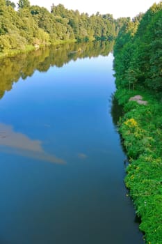 Beautiful river with a watery surface reflecting the trees growing on the shore and clouds in the sky. A good place to picnic and relax with friends.