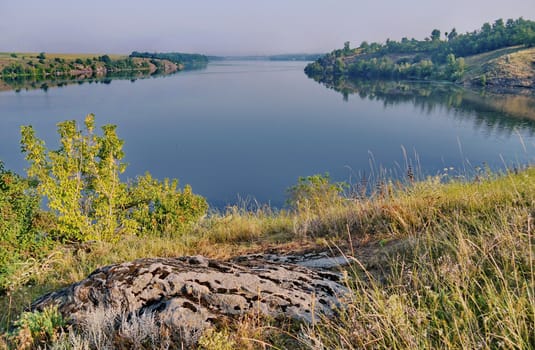 river surface against the background of rocky coasts with summer dry grass and green bushes under the gray sky. a place for rest, picnicking, fishing and tourism