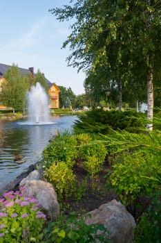 a fountain in the middle of an artificial lake near a wooden private house on the shore of which grow birches and coniferous bushes