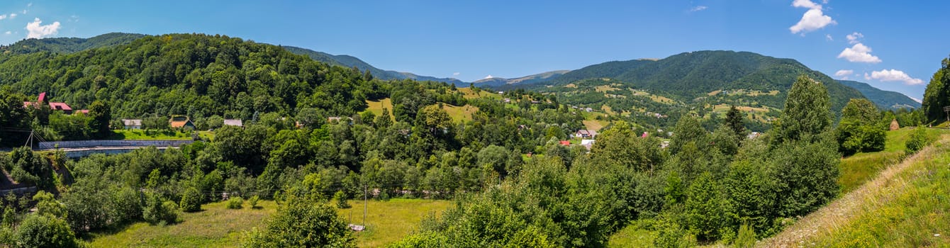 Beautiful panorama of the village stretched in a mountain valley and on the slopes covered with green trees under a cloudless summer sky.