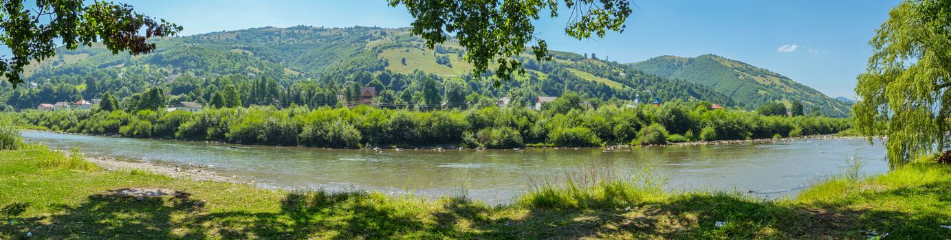 A beautiful panorama of the mountain river enveloping the shore with the visible roofs of the houses against the background of green trees and mountain slopes with a growing forest behind them.