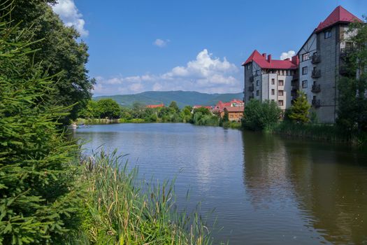 A small transparent mountain lake against the backdrop of the buildings of the hotel complex and green coniferous trees