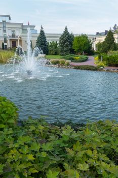 beautiful fountain in a pond surrounded by green on the background of a light mansion and a blue cloudless sky