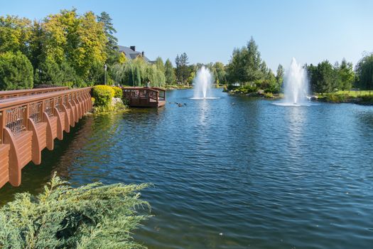 Excellent rest in the park with a pedestrian bridge pond with fountains and ducks. The beauty of a summer fair day against the blue sky.