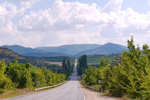 The path is between the trees and going to the mountains, under the sky with white clouds