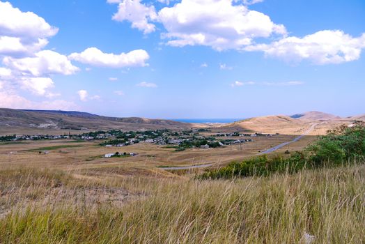 a landscape of vague gray hills and a settlement in a valley with a narrow strip of sea blue in the distance