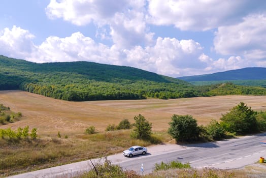 gray car on a road in the middle of a valley with dry grass and a dense green forest against a blue cloudy sky