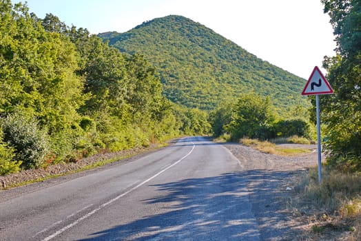 asphalt road resting on a mountain with a sign showing the turn with green trees on the roadside