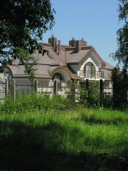 A solid house behind an iron fence with a fence with a small balcony with a growing green grass near the fence.