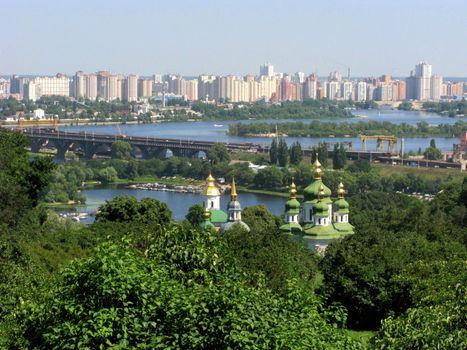 An excellent landscape with the tops of domes over green trees. A bridge going through a wide river and city blocks of high-rise buildings.