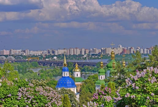 Beautiful golden domes of a church in leaves of blossoming lilacs against the background of a bridge across the river and houses of the city.