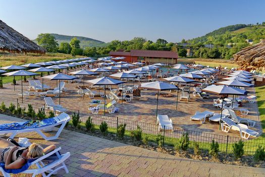 A poolside terrace is paved with sun loungers and umbrellas for holidaymakers against the background of high hills.