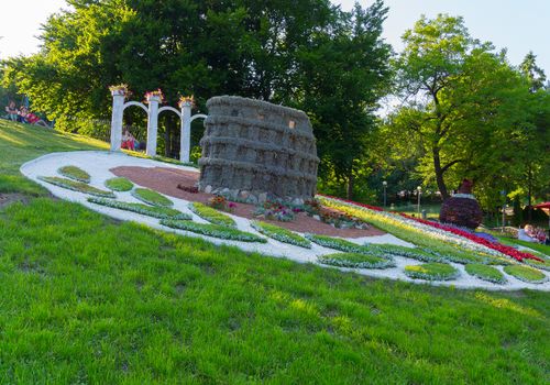 tourists in the park sitting on top of the slope contemplate a sculpture in the middle of the flowerbed made in the form of a fortress