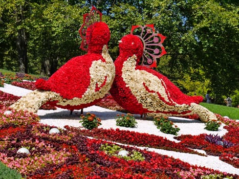 Silhouette of two large birds made of red and white flowers against the background of a beautiful flowerbed and deciduous trees.