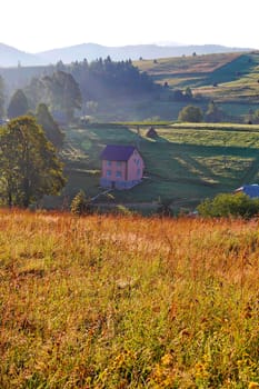 view from the top of the hill to a sunlit valley with a two-story house, fields and trees against a cloudless sky and mountains