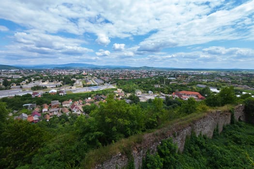 View of the roofs and houses of the modern city from behind the collapsed wall of an ancient castle overgrown with bushes and grass.