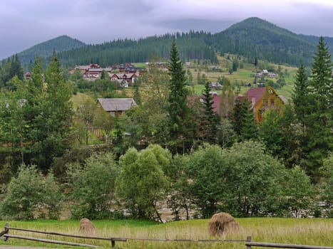 A view of the houses surrounded by trees and a few haystacks in the foreground