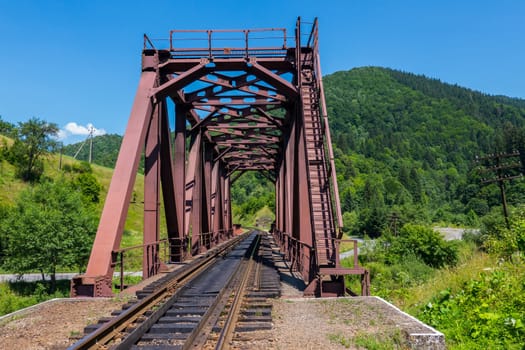 The old iron construction of the railway bridge against the background of the high green mountains