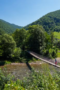 A tourist on a pedestrian bridge going over a shallow mountain river strewn with stones. A sunny summer day against the background of the surrounding nature.
