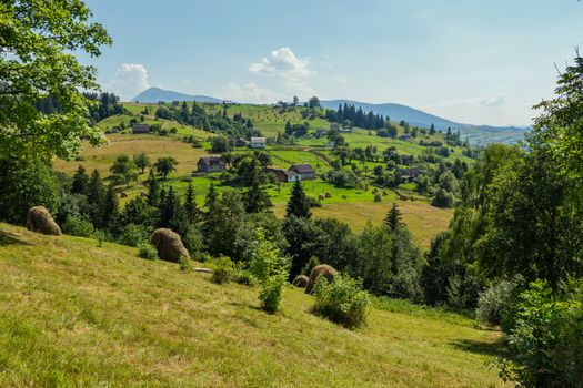 under the blue sky on the mountainside there are rural houses with well-kept fields, green trees, gathered in haystacks