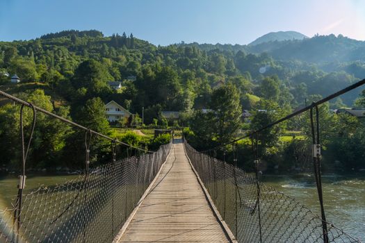 pedestrian bridge across a mountain river leading to rural houses drowning in the green of trees