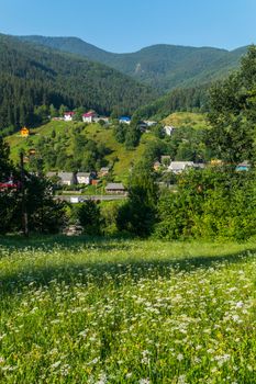Beautiful view from a green glade with flowers located on a hillside to the village houses and mountains located behind them.