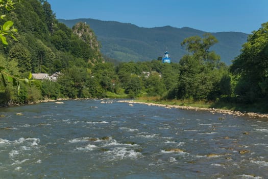 The swift mountain river against the background of the blue top of the church church, small houses and rocky mountains