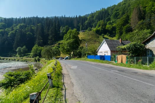 An asphalt road in a mountainous area with rural houses on one side and a pond on the other.