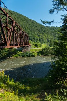 A railway bridge over a mountain river against the backdrop of a slope overgrown with green forest and a blue cloudless sky above it.