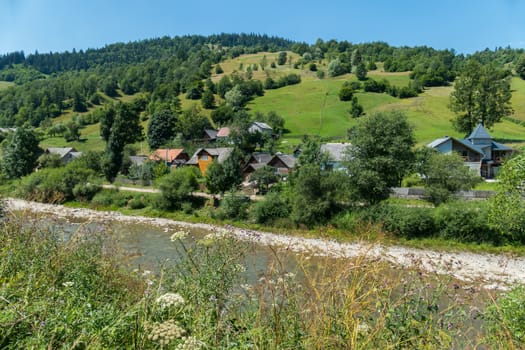 The village street with neat houses is located at the foot of the hill with green slopes and the current below the river.