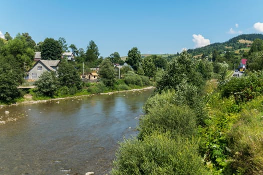a river flowing along the hidden among the green trees and bushes of private rural houses and forest-covered mountain peaks with a road far away in the distance under a blue cloudy sky. place of rest and tourism
