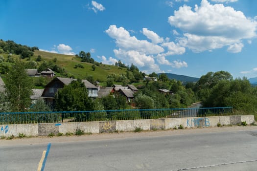 village on a green mountain slope behind an asphalt road on a bridge across a stormy river under a blue cloudy sky. place of rest and tourism