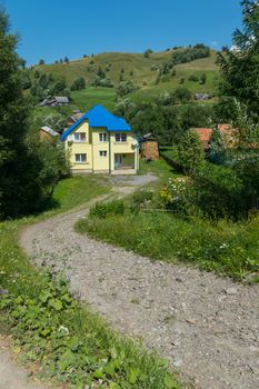 A mountain road leading to a yellow cottage with a blue roof against a high green slope