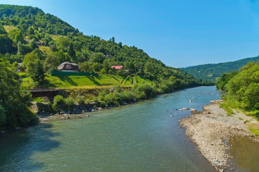 wide mountain river with tributaries and rocky shores near the village on the background of green hills