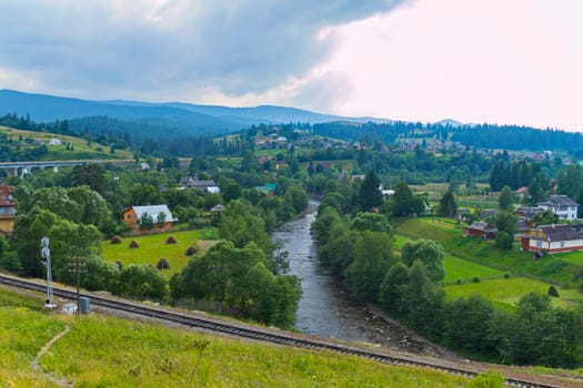 village on a green plain with a railway and a mountain river against a backdrop of distant peaks and a gray cloudy sky. place of rest and tourism