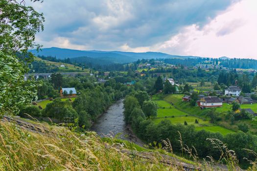 an exciting landscape on a mountain river with willows along the shores and a town on both sides of the stream against the background of the distant blue mountains