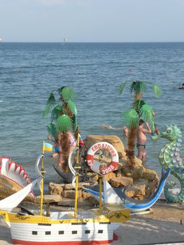 Beach beach with a ship layout on the beach with flags flying on the tops of the masts. Tourists are swimming in the waters of the sea stretching to the horizon line.