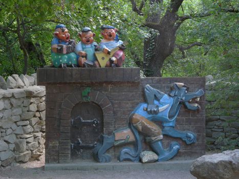 A merry sculpture in the park in the form of a wall with a wolf stalking with a knife and light-hearted pigs playing musical instruments and singing songs.