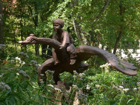 Sculpture of a boy sitting astride a flying goose against a background of green grass